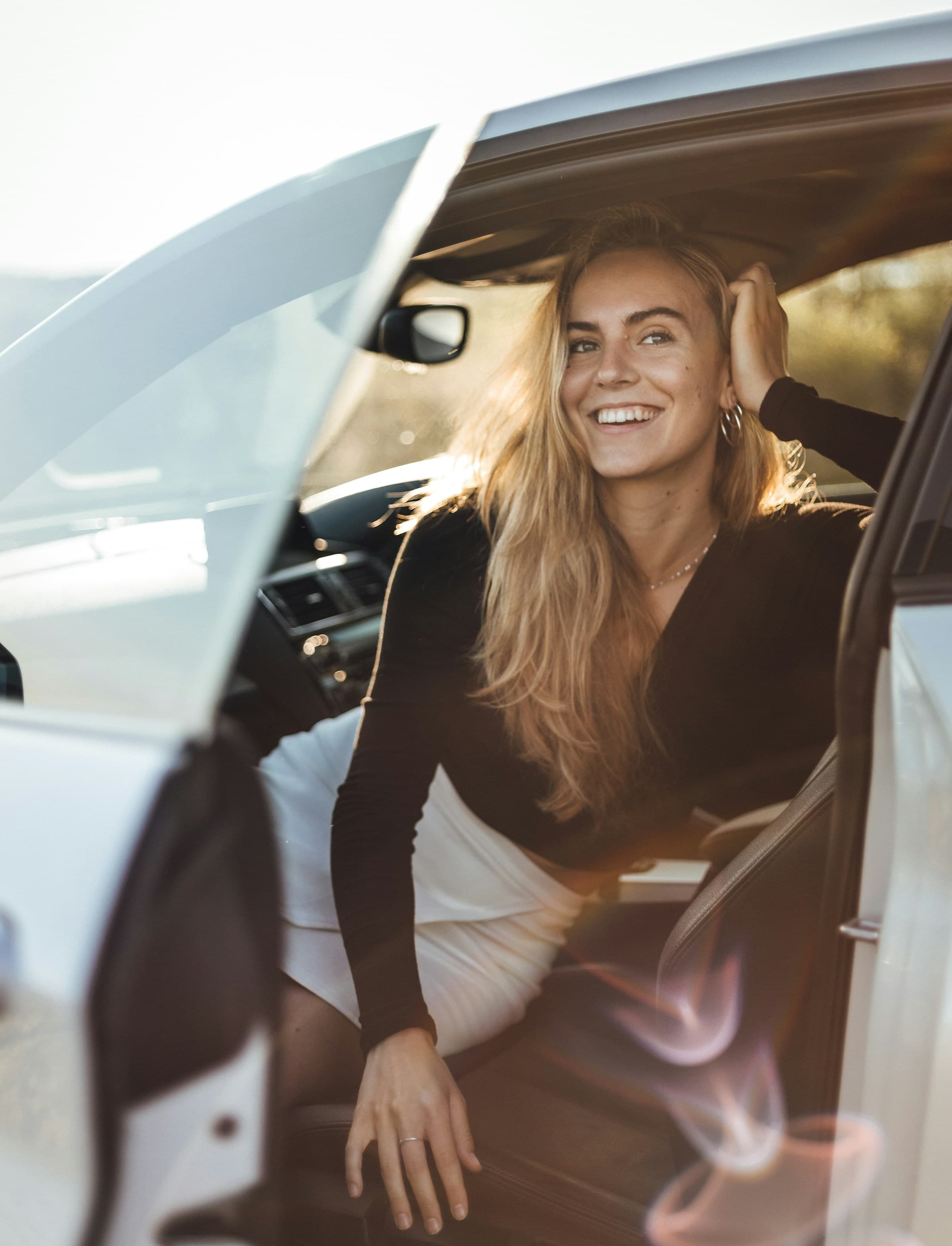 A woman laughing in the driver's seat of her car with the door open, in late-afternoon sun.